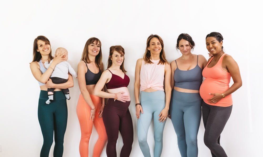 Groupe de 6 femmes "mixte" de tailles et formes différentes, certaines enceinte et autres jeunes mamans, dont une avec un bébé dans les bras, souriantes, faisants toutes parties de la communauté WoMum Studio