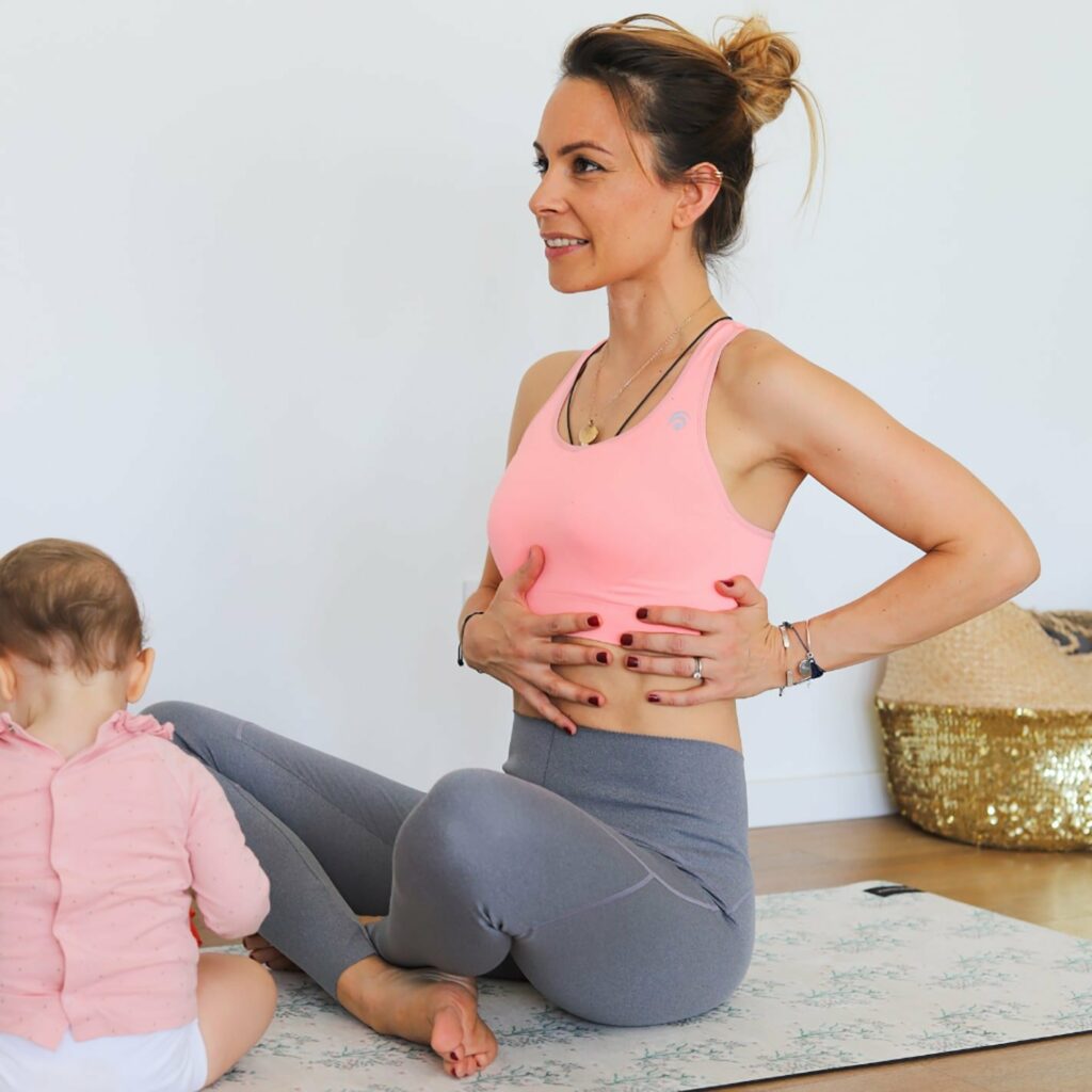 Femmes après son accouchement en train d'effectuer un des exercices de respiration sécurisés pour se reconnecter à son corps de femme après l'accouchement, assise en tailleur sur un tapis de yoga avec son bébé à coté d'elle
